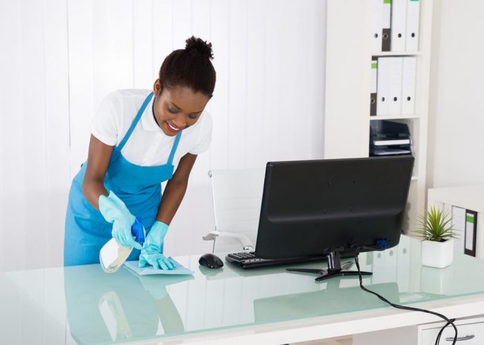 Happy Young African Female Janitor Cleaning Desk With Rag In Office